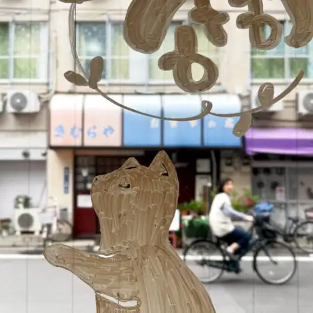 Through a window painted with a cat silhouette, we are looking at a suburban street in Itabashi, Tokyo, with a bicycle passing in front of an old restaurant.