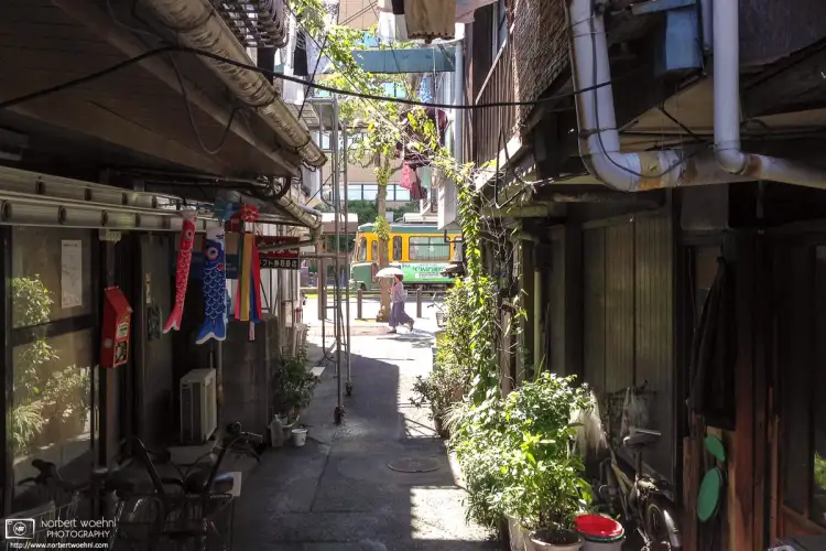 Through an alley of residential buildings in Kagoshima on the south western tip of the island of Kyūshū in Japan, I captured this view of a city tram and a woman carrying a parasol.