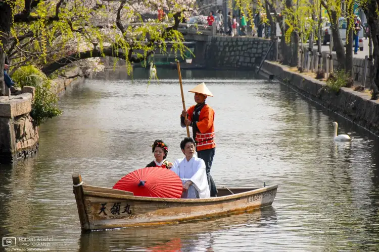 A man and a woman in traditional clothes and holding a paper umbrella are riding in a boat on the canal in the Bikan District of Kurashiki, Japan.
