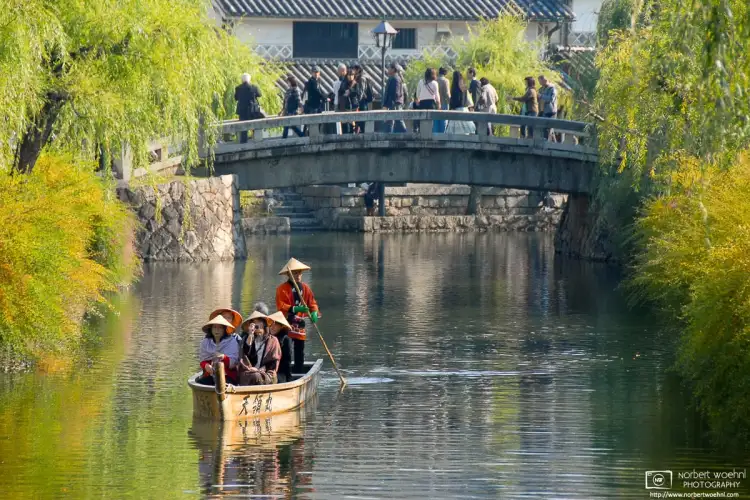 During early autumn season, visitors are enjoying a boat ride on a willow-lined canal in the historic Bikan district of Kurashiki in Okayama Prefecture, Japan.
