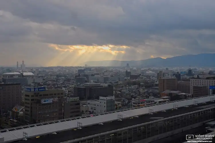 Rays of sunlight are breaking through the clouds to illuminate the southern part of central Kyoto. The pagoda of Toji Temple is visible towards the right side.