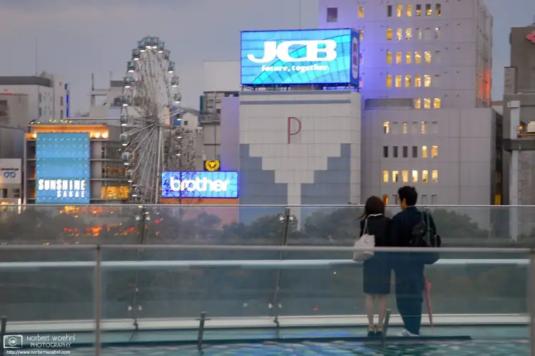 On the Spaceship Aqua rooftop of the Oasis 21 complex in the Sakae area of Nagoya, Japan, a young couple is looking towards some neon advertisements at dusk.