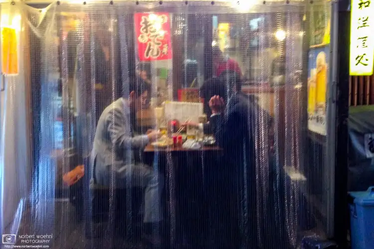 Two salarymen are enjoying a simple dinner seated outside a pub in the Nakano area of Tokyo, Japan, as seen through a plastic blind outside the shop.