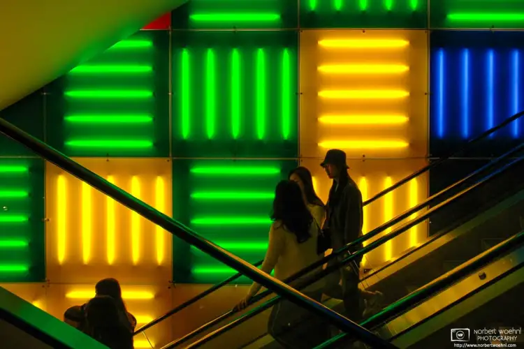 Against a striking background pattern of colorful neon lights, people are riding an escalator at a store on Times Square in Manhattan, New York.