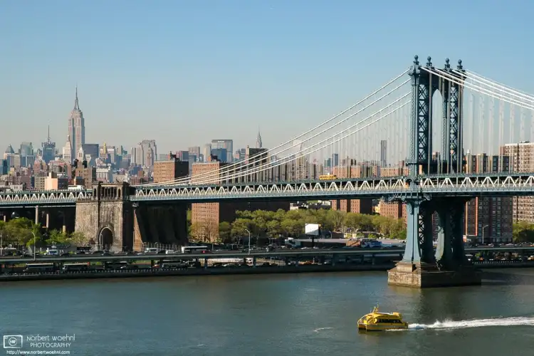 A water taxi is speeding along the East River in New York City. Manhattan Bridge and the Empire State Building are visible in the background.