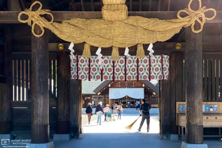 A uniformed man holding a brush is sweeping the area around the entrance to Hokkaido Jingu, a shrine in Sapporo, Japan.