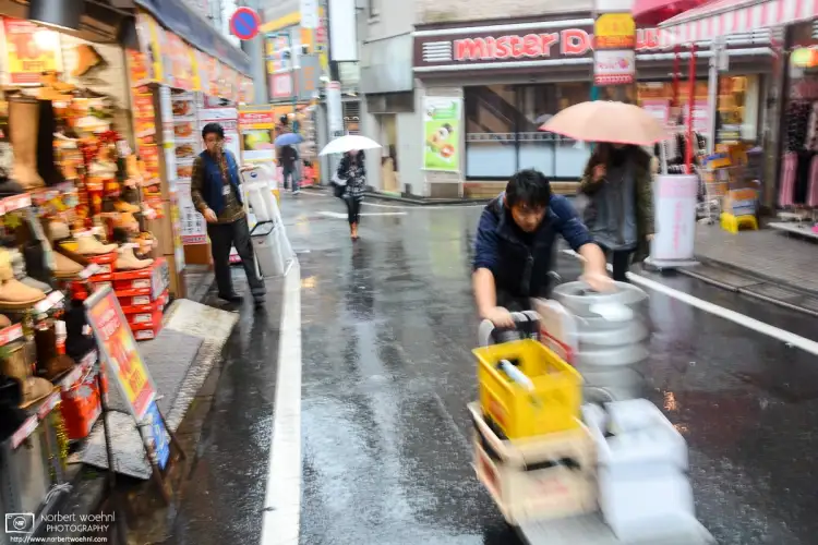 During pouring rain, a delivery person is pushing a cart with beverage containers around Shimokitazawa in Tokyo, Japan.