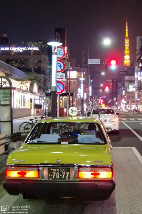 Outside Hamamatsucho Station in Tokyo, Japan, an old-school taxi looks ready for a ride that is headed towards the illuminated Tokyo Tower in the background.
