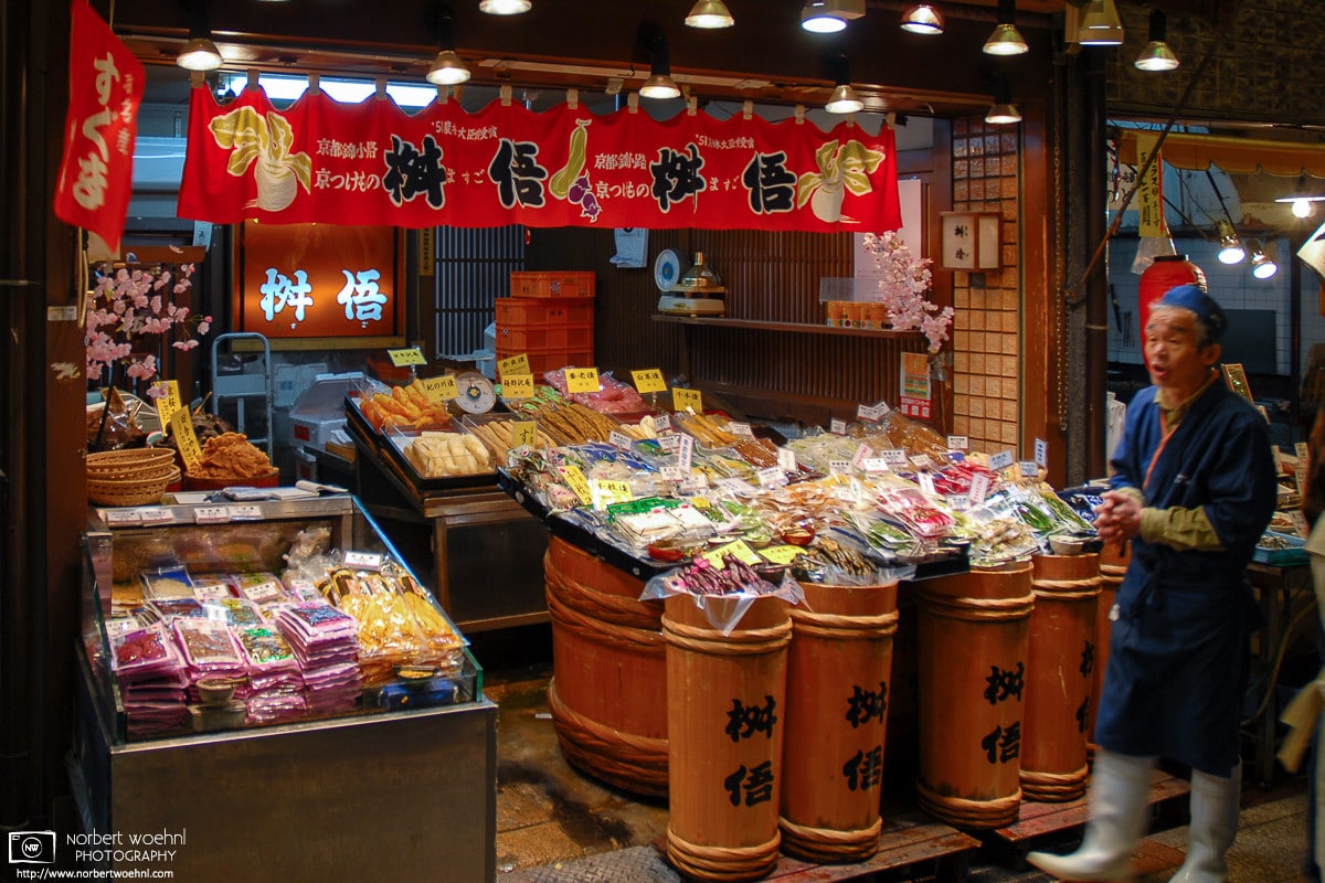 At Nishiki Market in Kyoto, Japan, a shop owner is seen outside his shop selling a variety of pickled vegetables.
